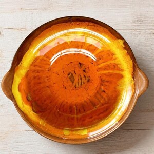 May include: A round ceramic baking dish featuring a vibrant gradient glaze that transitions from a deep brown rim to a bright yellow mid-section and a rich orange center. The dish has two small integrated handles on opposite sides.