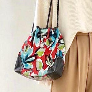 May include: A drawstring bucket bag featuring a vibrant red, light blue, and green floral pattern. The bag includes black leather-like corner reinforcements, thin braided black shoulder straps, and a silver-toned metal charm attached to the drawstring.