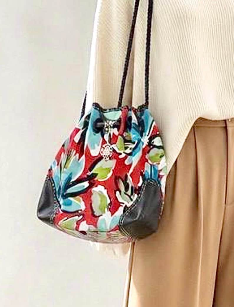 May include: A drawstring bucket bag featuring a vibrant red, light blue, and green floral pattern. The bag includes black leather-like corner reinforcements, thin braided black shoulder straps, and a silver-toned metal charm attached to the drawstring.