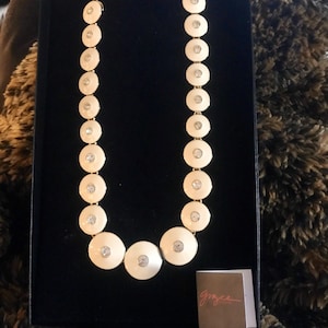 May include: A white and gold necklace with round, white, enamel-like pendants. Each pendant has a clear rhinestone in the center. The necklace is displayed in a black box with a small white tag that says "Grace".