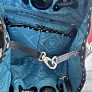 May include: The interior of a bag featuring a bright blue lining with a repeating circular logo pattern. It includes a black zippered pocket, a black leather strap with a silver metal lobster clasp, and black leather trim along the top edge.
