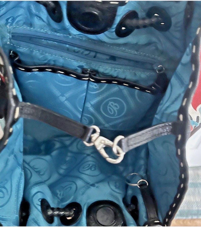 May include: The interior of a bag featuring a bright blue lining with a repeating circular logo pattern. It includes a black zippered pocket, a black leather strap with a silver metal lobster clasp, and black leather trim along the top edge.