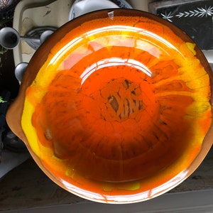 May include: A large, round, orange and yellow ceramic bowl with a glazed finish. The bowl has a unique swirl pattern and a slightly raised rim.