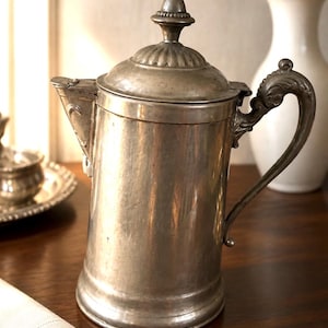 May include: A metallic silver-toned coffee pot with a cylindrical body, a hinged lid featuring a decorative finial, a curved handle with ornate detailing, and a short spout. The surface has a matte finish with visible signs of wear and texture.