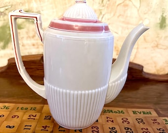 May include: White ceramic coffee pot with a pink and red trim around the lid. The pot has a fluted design and a handle with a red trim. The pot is sitting on a wooden surface with a game board.