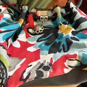 May include: A bucket bag featuring a canvas body with a vibrant floral print in red, turquoise, black, and yellow. The bag includes black braided leather handles, a black leather base, and a silver-toned metal charm attached to the drawstring cord.