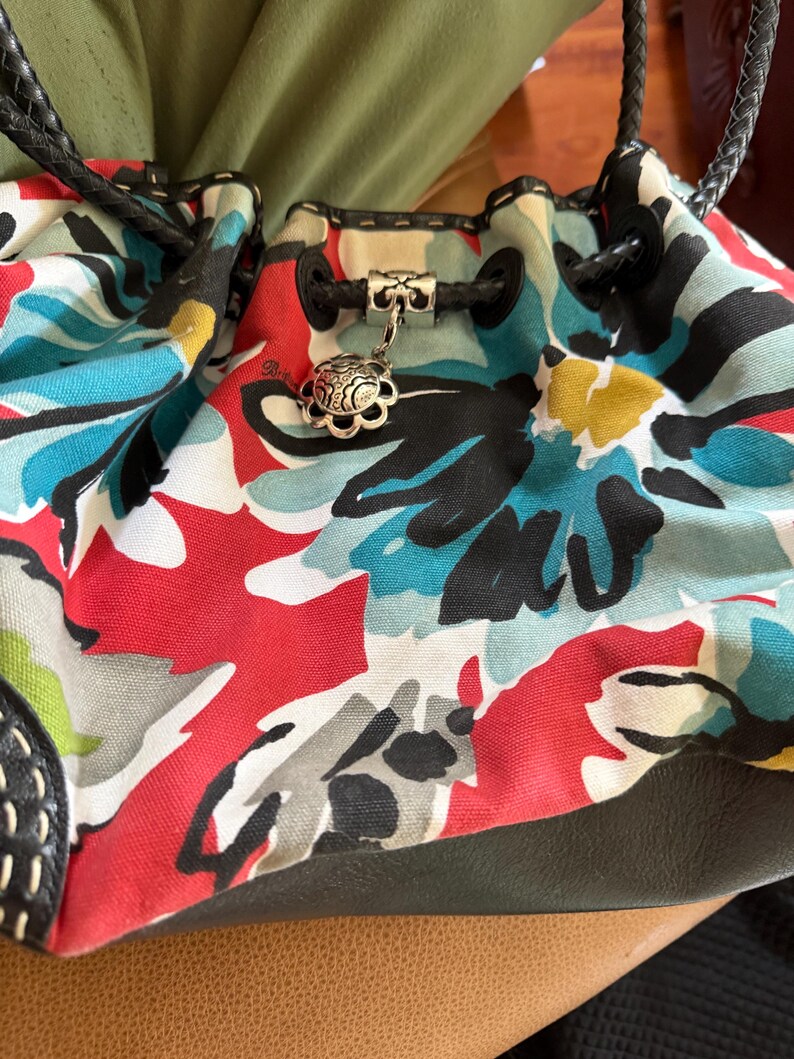 May include: A bucket bag featuring a canvas body with a vibrant floral print in red, turquoise, black, and yellow. The bag includes black braided leather handles, a black leather base, and a silver-toned metal charm attached to the drawstring cord.