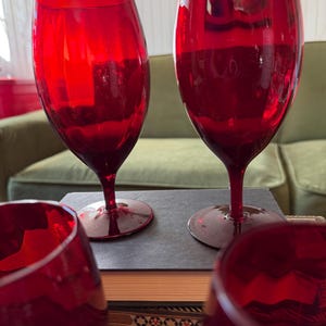 May include: Four ruby red wine glasses with long stems are arranged on a dark surface. The glasses are made of transparent glass and reflect light. The background includes a green sofa and decorative elements.