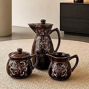 May include: A dark brown ceramic tea set consisting of a lidded pitcher, a lidded sugar bowl, and a creamer. Each piece features a hand-painted floral pattern in white and light pink, with a glossy finish and matching curved handles on each item.