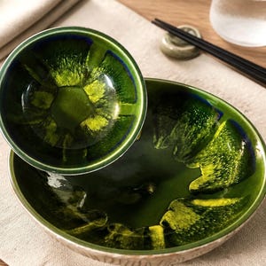 May include: A set of two ceramic bowls featuring a glossy, reactive glaze in shades of deep forest green, bright lime, and dark blue. The bowls have a smooth, reflective interior surface with a textured, light-colored exterior rim and base.