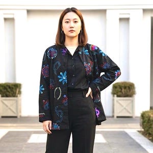 May include: A black long-sleeved jacket featuring colorful embroidered patterns in shades of blue, purple, and red. The jacket has a collarless neckline and a button-down front, worn over a solid black button-down shirt and matching black trousers.