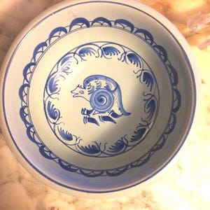 May include: A blue and white ceramic bowl with a stylized bird design. The bowl has a blue rim and a blue and white floral pattern around the bird.