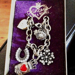 May include: A silver charm bracelet with a purple velvet box. The bracelet features charms including a heart, horseshoe, snowflake, and a crown with a red heart. The box has the text "Princess Vera Wang" on the lid.