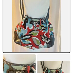 May include: A hobo-style handbag featuring a vibrant red base with a large-scale floral print in shades of blue, white, and green. The bag includes dark brown leather trim, braided shoulder straps, a drawstring closure, and a silver-toned metal charm.