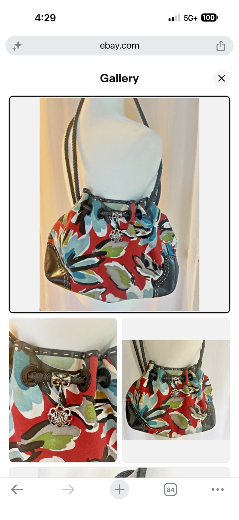 May include: A hobo-style handbag featuring a vibrant red base with a large-scale floral print in shades of blue, white, and green. The bag includes dark brown leather trim, braided shoulder straps, a drawstring closure, and a silver-toned metal charm.