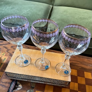 May include: Three clear wine glasses with a decorative purple fleur-de-lis pattern around the rim and gold trim. The glasses sit on a wooden box with a decorative inlay. Each glass has a blue sticker on the base.