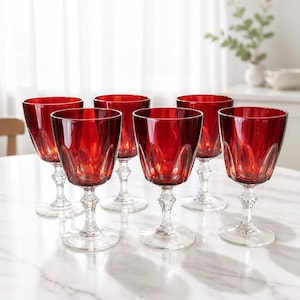 May include: A set of six ruby red glass goblets with clear stems and bases. Each goblet features a wide bowl with a vertical ribbed pattern at the base, transitioning into a smooth rim. The stems have a faceted, decorative design.