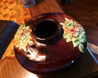 May include: A brown ceramic vase with a glossy finish. The vase features a dark opening and is adorned with raised floral designs in shades of yellow, pink, and green. The vase is a decorative item.
