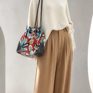 May include: A cream-colored ribbed knit sweater paired with tan wide-leg trousers and a drawstring bucket bag. The bag features a vibrant red, blue, and green floral pattern with black leather accents and thin, braided black shoulder straps.