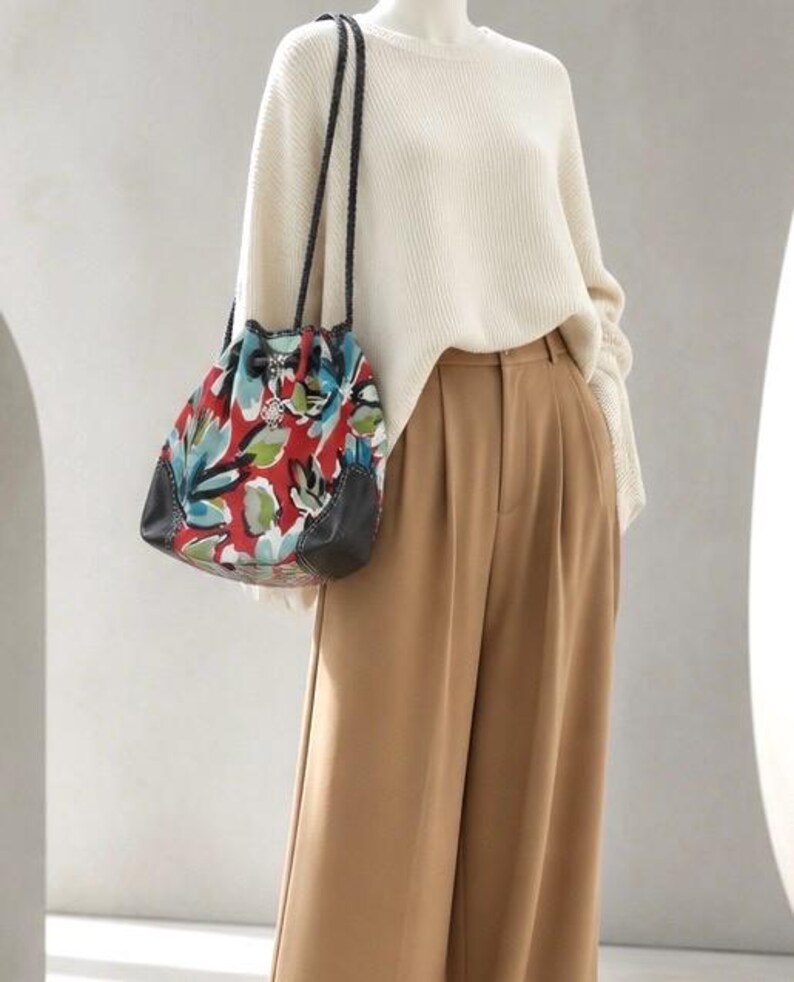 May include: A cream-colored ribbed knit sweater paired with tan wide-leg trousers and a drawstring bucket bag. The bag features a vibrant red, blue, and green floral pattern with black leather accents and thin, braided black shoulder straps.