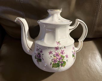 Può includere: Una teiera bianca con un design floreale con fiori viola e foglie verdi. La teiera ha un bordo dorato attorno al coperchio e al beccuccio.