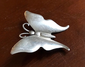 May include: A silver butterfly brooch with textured wings. The butterfly is in flight with its wings spread.