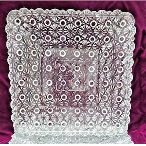 May include: A square clear glass plate featuring a repeating geometric pattern of raised starbursts and small circular indentations. The edges of the plate are scalloped with a decorative border, and the center has a smooth, recessed square section.