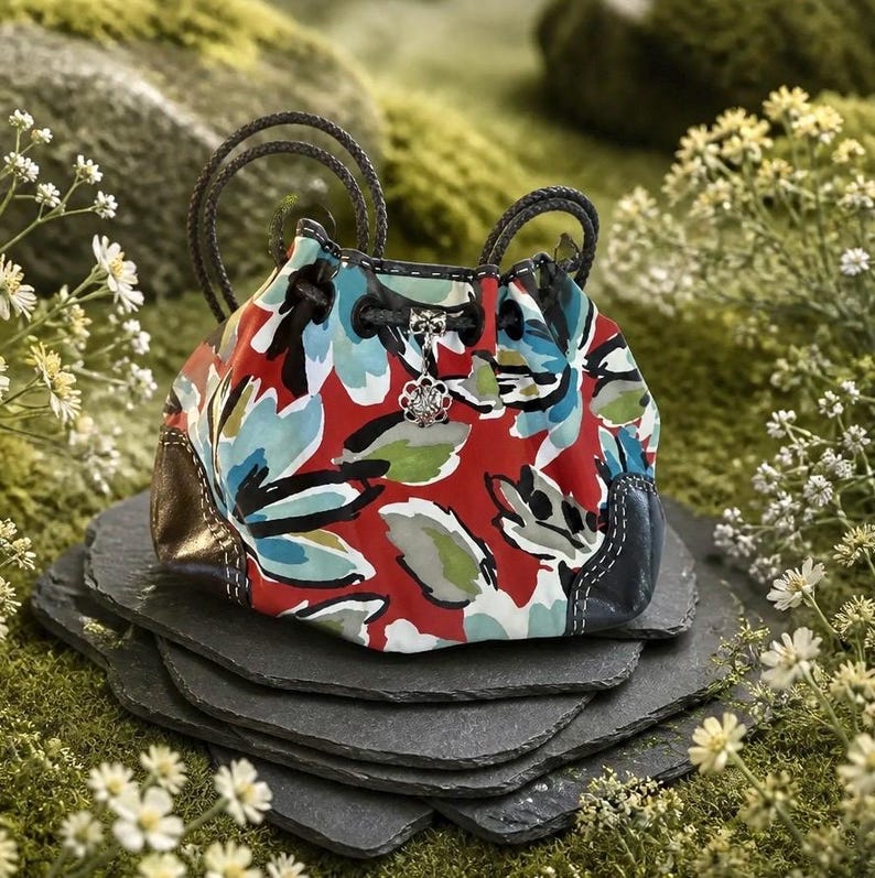 May include: A drawstring pouch featuring a vibrant floral pattern in red, light blue, and green. The bag has two braided dark handles, a metallic silver flower-shaped charm attached to the drawstring, and metallic bronze corner accents.