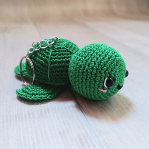 Op de afbeelding: Een handgemaakte groene gehaakte schildpad sleutelhanger. De schildpad heeft een ronde kop met zwarte ogen en een kleine roze mond. De sleutelhanger is bevestigd aan de rug van de schildpad met een zilveren metalen ring.