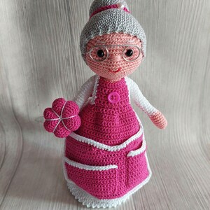 Può includere: Una bambola all'uncinetto che indossa un vestito rosa con un grembiule bianco e un cappello bianco all'uncinetto. La bambola tiene un fiore rosa all'uncinetto.
