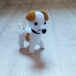 Può includere: Un giocattolo per cani in crochet bianco con orecchie marroni e occhi neri. Il cane è in piedi su una superficie di legno chiaro.
