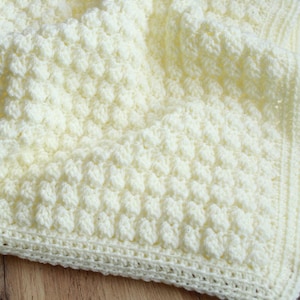 May include: A white crocheted baby blanket with a textured, puffy stitch pattern. The blanket has a simple border.