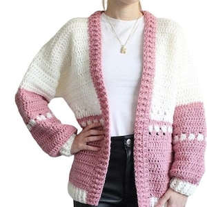 May include: A pink and white crocheted cardigan with long sleeves and a v-neck. The cardigan is made with chunky yarn and has a relaxed fit.