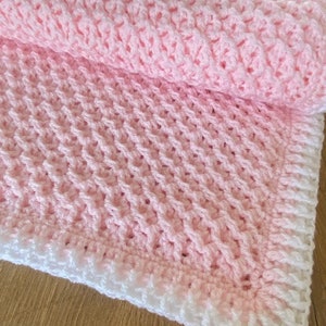 Op de afbeelding: Een roze gehaakte babydeken met een witte rand. De deken is gemaakt met een textuurpatroon.