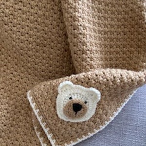 May include: A brown crocheted baby blanket with a white crocheted border. A white crocheted bear applique is in the lower right corner of the blanket.