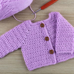 Puede incluir: Un cárdigan de bebé de ganchillo de color lavanda con botones de madera.