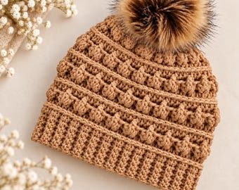 May include: A tan crochet beanie featuring a ribbed brim and a textured pattern of raised bobble stitches across the body. The hat is topped with a large, fluffy, multi-toned brown faux fur pom-pom that adds volume to the overall design.