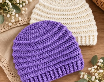 May include: Two crochet beanies, one in a vibrant purple and one in a soft cream color. Both hats feature a textured, ribbed stitch pattern with a thick, folded brim at the base. The beanies are crafted with a consistent yarn weight and stitch.