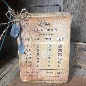 May include: A rectangular kitchen conversion chart with a handle, featuring measurements for cups, ounces, tablespoons, and teaspoons. The chart has a rustic, vintage aesthetic with a metal spoon and tag tied with twine.