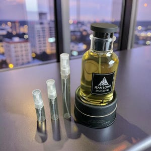 May include: A perfume bottle with a black cap and a label that reads "JEAN LOWE immortel" sits on a black circular base. Three small, clear spray bottles are next to it. The perfume is a light amber color.