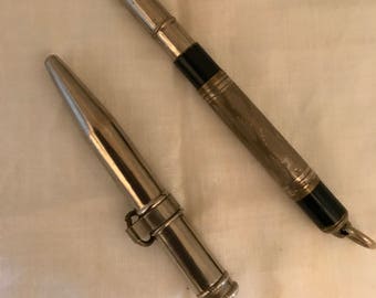 May include: Two vintage silver-toned metal objects. One is a pen with black accents and a ring at the end. The other is a pointed object with a hinged ring. Both objects are on a white surface with a blue floral accent.