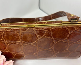 May include: A vintage brown handbag with a textured, crocodile-like pattern. The bag has a gold-toned clasp and handle attachments. The handle is a darker brown leather. A classic accessory.