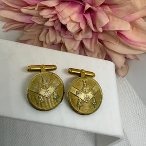 Könnte beinhalten: Ein Paar runde, goldfarbene Manschettenknöpfe mit einer strukturierten Oberfläche, einem eingravierten Chevron-Muster und kleinen glockenförmigen Motiven. Die Manschettenknöpfe haben ein poliertes Gold-Finish und einen schwenkbaren Knebelverschluss.