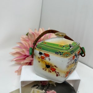 Op de afbeelding: Een vintage, vierkante keramische container met een deksel en een geweven handvat. De container is wit met kleurrijke bloemen- en vogelontwerpen, groene accenten en gouden rand. Het handvat is bruin en bevestigd met groene accenten.