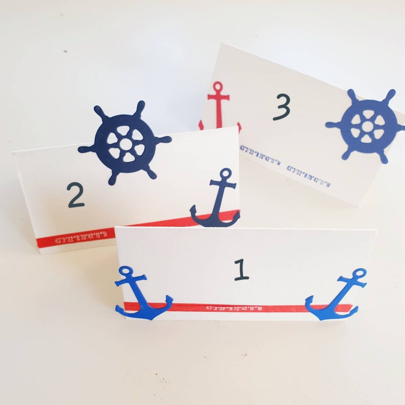 Peut inclure: Trois cartes de place avec des th&egrave;mes nautiques. Chaque carte a un num&eacute;ro, une ancre bleue et un volant bleu. Les cartes ont une bande rouge et blanche en bas.