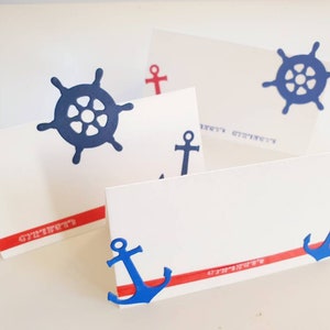 Peut inclure: Quatre cartes de place blanches avec des d&eacute;corations nautiques rouges et bleues. Les d&eacute;corations comprennent des ancres et des volants. Les cartes de place ont une bande rouge avec le mot "Strawberry" imprim&eacute; dessus.