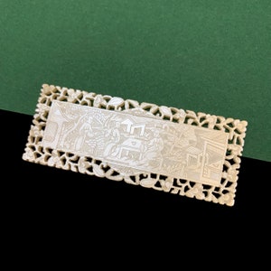 Puede incluir: Una pieza decorativa rectangular blanca con un diseño tallado intrincado. El diseño presenta una escena de personas en un entorno de jardín con edificios en el fondo.