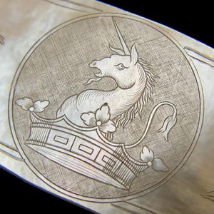 May include: Detailed silver engraving of a unicorn with a crown, set within a circular frame. The design features intricate linework, floral accents on the crown, and a cross-hatch background pattern. A decorative element.