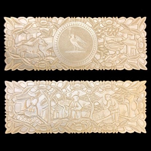 Puede incluir: Dos piezas rectangulares de nácar están talladas con diseños intrincados. La pieza superior presenta un pájaro en un círculo, mientras que la pieza inferior representa personas en un paisaje.