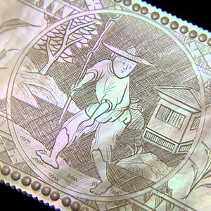 May include: Close-up of a detailed, engraved artwork depicting a figure in traditional attire, holding a staff and walking near a building and trees. The artwork is set within a circular frame with a decorative border, showcasing intricate line work and a pearlescent sheen.